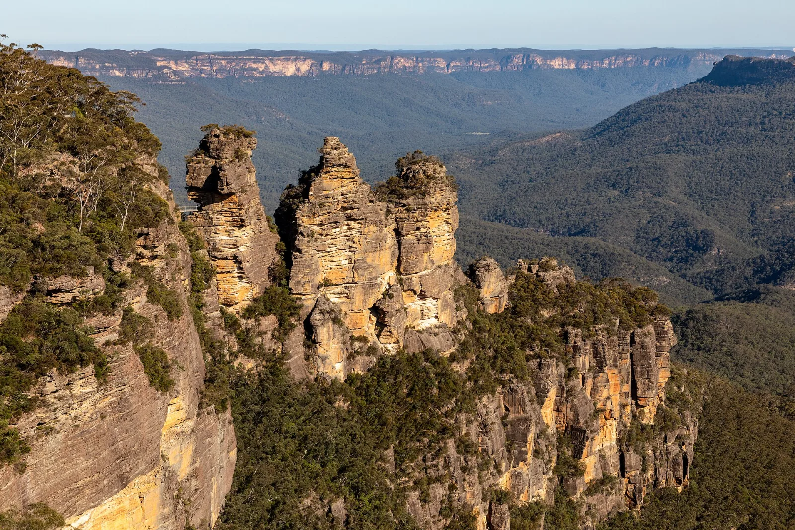 Three Sisters, Blue Mountains National Park, New South Wales, AustraliaThe Blue Mountains National Park is located west of Sydney and is one of Australia’s most famous natural areas. Its history is closely linked to Aboriginal culture, particularly that of the Gundungurra and Darug peoples, who have inhabited the area for tens of thousands of years and have a spiritual connection to the landscape.For European settlers, the rugged sandstone plateaus were long considered an insurmountable obstacle. It was not until 1813 that Gregory Blaxland, William Lawson, and William Charles Wentworth succeeded in crossing the mountains, opening the way for the development of the Australian hinterland.In the 19th century, parts of the region were used for logging and mining, while at the same time its scenic appeal was discovered. In 1959, the Blue Mountains National Park was officially established to protect the unique eucalyptus vegetation, deep gorges, and waterfalls.In 2000, UNESCO declared the area part of the Greater Blue Mountains Area a World Heritage Site. Today, the park stands for nature conservation, tourism, and recognition of Australia’s indigenous cultural history.